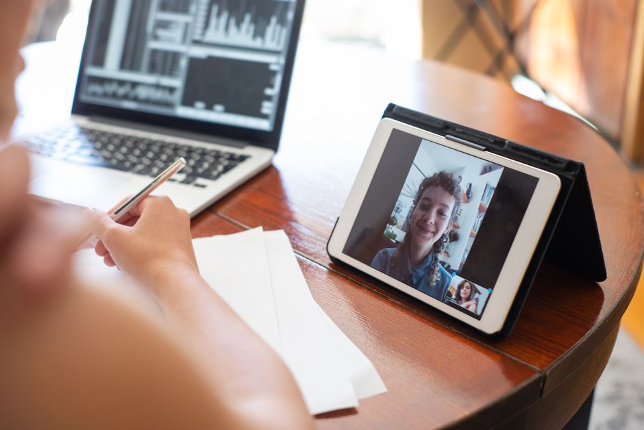 Uma pessoa em uma chamada de vídeo usando um tablet, com um laptop exibindo dados nas proximidades.