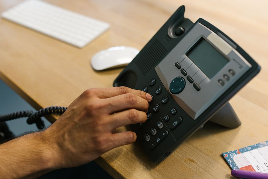 Uma mão discando um telefone em uma mesa de madeira. Ambiente de escritório com teclado e planner.